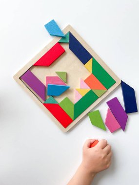 Childs el, beyaz arka planda çok renkli ahşap mozaik koleksiyonu yapıyor. Çocuk renkli bir tangram çözüyor. Beyaz arkaplanda renkli geometrik şekillerin karesi.