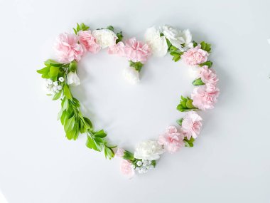 Beyaz arka planda pembe ve beyaz karanfillerden, yeşil yapraklardan, çingenelerden oluşan bir kalp şeklinde çerçevelenmiş. Düz yerde, üst tarafta. Bahar arka planı. Düğün, kartlar ve davetiyeler için uygun