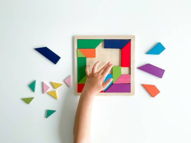 Childs el, beyaz arka planda çok renkli ahşap mozaik koleksiyonu yapıyor. Çocuk renkli bir tangram çözüyor. Beyaz arkaplanda renkli geometrik şekillerin karesi.