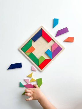 Childs el, beyaz arka planda çok renkli ahşap mozaik koleksiyonu yapıyor. Çocuk renkli bir tangram çözüyor. Beyaz arkaplanda renkli geometrik şekillerin karesi.
