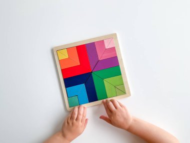 Childs el, beyaz arka planda çok renkli ahşap mozaik koleksiyonu yapıyor. Çocuk renkli bir tangram çözüyor. Beyaz arkaplanda renkli geometrik şekillerin karesi.
