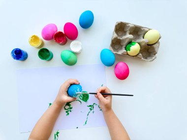 Çocuklar Paskalya yumurtalarını renkli yumurtalar ve beyaz masadaki boya kavanozlarıyla çevrili fırçayla boyarlar. Yaratıcı tatil aktiviteleri. Yüksek kalite fotoğraf