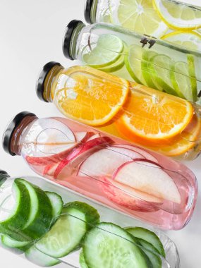 Beyaz zemin üzerinde salatalık, elma, portakal ve limon dilimleri olan detoks içeceklerle dolu cam şişeler. Dikey yönelim, tasarım ve baskı için canlandırıcı içecek konsepti. Yüksek kalite fotoğraf