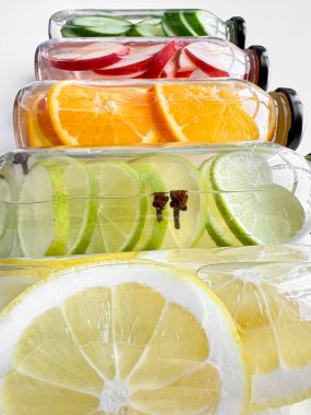 Beyaz zemin üzerinde salatalık, elma, portakal ve limon dilimleri olan detoks içeceklerle dolu cam şişeler. Dikey yönelim, tasarım ve baskı için canlandırıcı içecek konsepti. Yüksek kalite fotoğraf