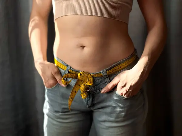 stock image Close up of woman faceless with tape measure in jeans instead of belt, standing with her hands in her pockets. Weight loss, fitness and body measurement concept. Lifestyle photo. High quality photo