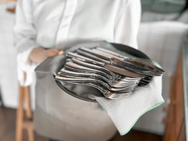 stock image Waiter in white shirt holding black tray with neatly arranged silverware including forks and knives on grey napkin, ready for table setting with blurred background of restaurant interior. High quality