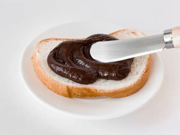 Taze ekmeğe sürülmüş kremalı çikolata ve beyaz tabakta tereyağı bıçağı..