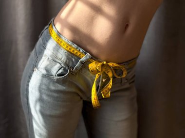 Close up of the naked torso of faceless woman in jeans with yellow measuring tape instead of belt on neutral background. Weight loss, fitness or body measurement concept. Lifestyle photo. High quality clipart