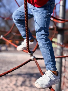 Çocuk, oyun parkında kot pantolon ve spor ayakkabılarla ipe tırmanıyor..