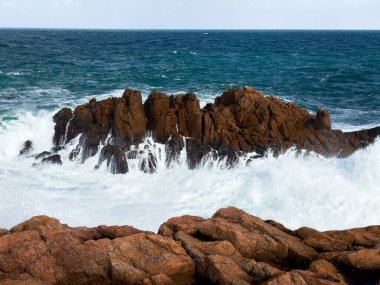 Dalgalar mavi okyanus gökyüzü ile kayalık kıyı şeridine çarpıyor.