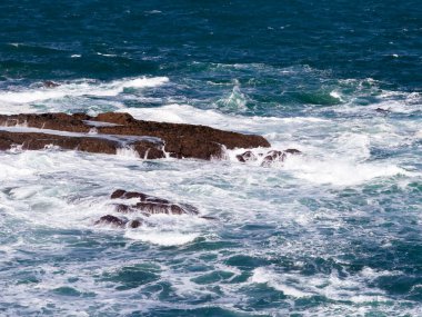 Dalgalar açık mavi gökyüzünün altında kayalık deniz kıyısına çarpıyor..