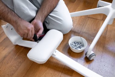 Erkek yetişkin beyaz ofis sandalyesini ahşap zeminde aletler ve vidayla birleştiriyor..