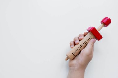 Beyaz arka planda kırmızı aksanlı ahşap oyuncak şırınga tutan çocuk eli.