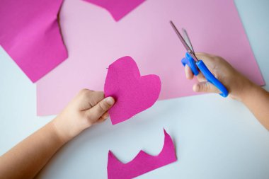 Pembe kağıt üzerinde mavi makasla pembe kalp şeklini kesen çocuk.