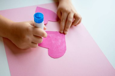 Çocuk pembe kağıt ve yapıştırıcıyla kalp şeklinde el sanatları yapıyor..