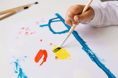 Beyaz kağıt üzerinde boya fırçasıyla renkli ve soyut bir resim yapan çocuk.