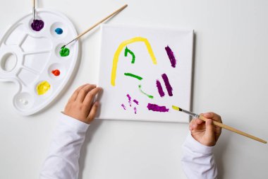 Çocuk renk paleti ve fırçalarla tuvale soyut resim çiziyor.