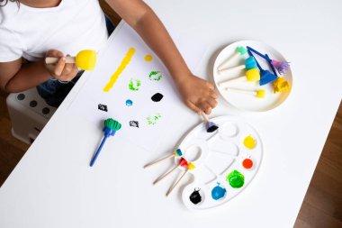 Child engaging in creative painting with colorful art supplies on white table. High quality.