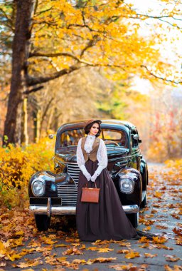 Eski moda elbiseli, dantel bluzlu ve şapkalı güzel bir kadın sonbahar arka planında retro kahverengi arabanın yanında duruyor. Eldiven giymiş zarif bir bayan. Sıcak sanat eserleri..