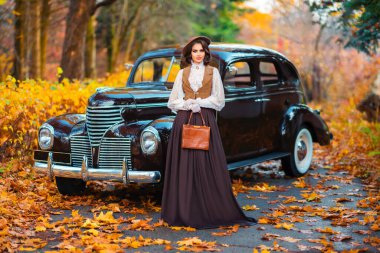 Eski moda elbiseli, dantel bluzlu ve şapkalı güzel bir kadın sonbahar arka planında retro kahverengi arabanın yanında duruyor. Eldiven giymiş zarif bir bayan. Sıcak sanat eserleri..