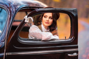 Eski moda elbiseli, dantel bluzlu ve peçeli güzel bir kadın sonbahar arka planında retro kahverengi arabada oturuyor. Eldiven giymiş zarif bir bayan. Sıcak sanat eserleri..