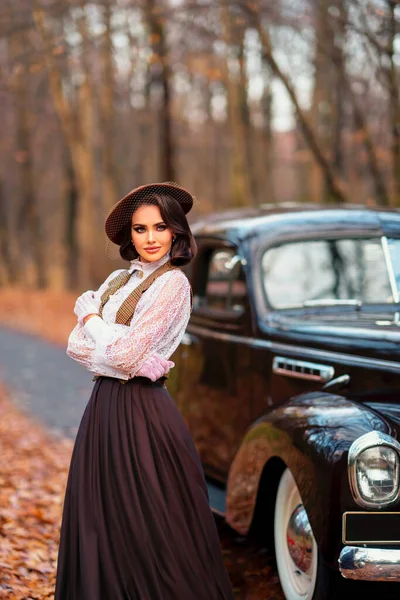 在秋天的背景下 穿着老式服装 花边衬衫 头戴面纱的漂亮女性站在复古棕色轿车旁边 穿着手套在大自然上摆姿势的优雅女士 温暖的艺术作品 — 图库照片