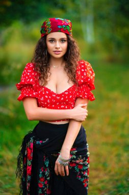 Pretty brunette curly girl in red gipsy costume and accessories posing on nature with green background and bonfire. Warm art work with queen. clipart