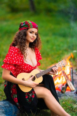 Pretty brunette curly girl in red gipsy costume and accessories posing on nature green background. Queen playing on guitar. clipart