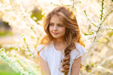 Portrait of beautiful young blonde curly girl with blue eyes posing on spring nature with white blooming trees.Sunny positive colorful photo with amazing teenage model. Light in hair.  
