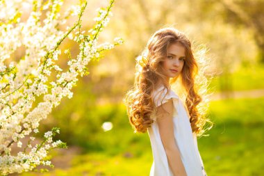 Portrait of beautiful young blonde curly girl with blue eyes posing on spring nature with white blooming trees.Sunny positive colorful photo with amazing teenage model. Light in hair.  