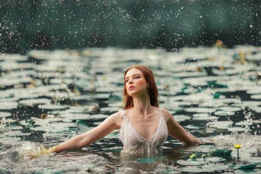 Beautiful red haired girl in white dress posing in river with water lilies. Fairytale story about  ophelia .Warm art work