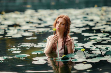 Beautiful red haired girl in white dress posing in river with water lilies. Fairytale story about  ophelia .Warm art work