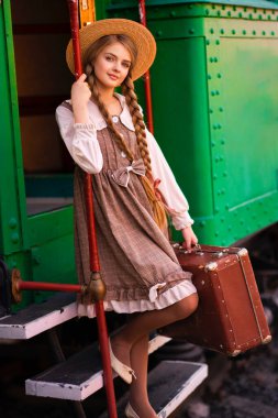 Yeşil trenin yanındaki istasyon platformunda dikilen klasik elbiseli ve hasır şapkalı güzel sarışın kızın portresi. Gülümseyen zarif, mavi gözlü hoş bayan. Gezgin hakkında sanat çalışması.