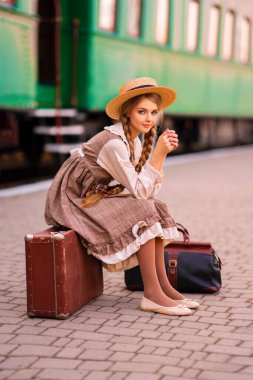 Yeşil trenin yanındaki istasyon platformunda dikilen klasik elbiseli ve hasır şapkalı güzel sarışın kızın portresi. Gülümseyen zarif, mavi gözlü hoş bayan. Gezgin hakkında sanat çalışması.