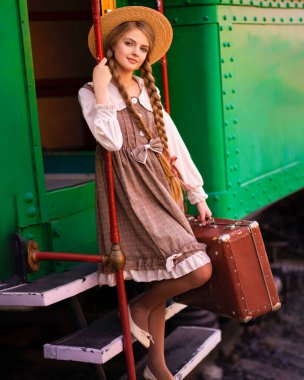 Yeşil trenin yanındaki istasyon platformunda dikilen klasik elbiseli ve hasır şapkalı güzel sarışın kızın portresi. Gülümseyen zarif, mavi gözlü hoş bayan. Gezgin hakkında sanat çalışması.