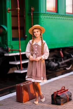 Yeşil trenin yanındaki istasyon platformunda dikilen klasik elbiseli ve hasır şapkalı güzel sarışın kızın portresi. Gülümseyen zarif, mavi gözlü hoş bayan. Gezgin hakkında sanat çalışması.