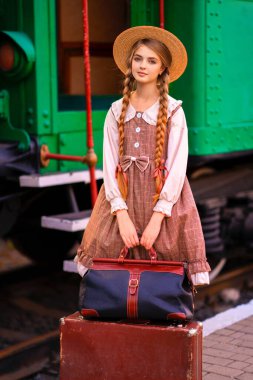 Eski moda elbiseli ve hasır şapkalı güzel sarışın kızın portresi yeşil trenin yanındaki istasyonda duruyor. Gülümseyen zarif, mavi gözlü hoş bayan. Gezgin hakkında sanat çalışması.