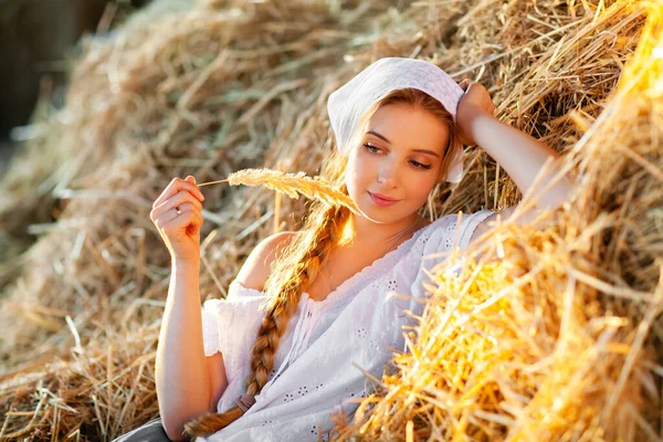 Beyaz kırsal kıyafetlerle örülmüş saçları olan güzel sarışın kız pipetin üzerinde oturuyor. Çiftlik hayatı. Çiftlikte poz veren genç bir model. Sıcak sanat eseri..