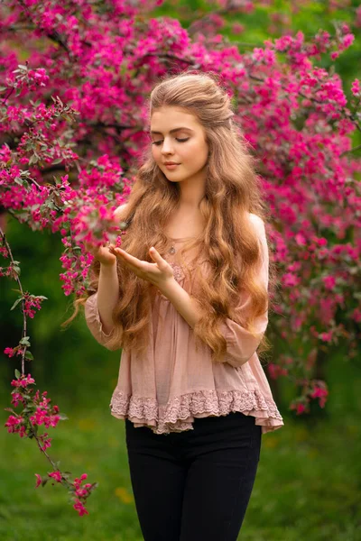 ピンクの花の近くの春の公園に立ってヴィンテージレースブラウスと緑のマントで長い巻き毛を持つかなり若いブロンドの女の子 優しさロマンチックなモデルポーズとカメラを見て — ストック写真