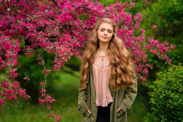 美丽的金发姑娘 卷曲长发 穿着老式花边衬衫和绿色斗篷 站在靠近粉色花朵的春天公园里 温柔浪漫的模特摆出姿势 看着镜头 — 图库照片
