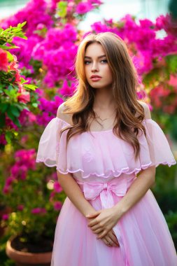 Renkli çiçeklerin yanında duran pembe klasik elbiseli ve hasır şapkalı güzel kız. Romantik bir kadının sanat eseri. Sevimli bir model, poz veriyor ve kameraya bakıyor..