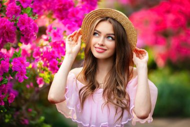 Renkli çiçeklerin yanında duran pembe klasik elbiseli ve hasır şapkalı güzel kız. Romantik bir kadının sanat eseri. Sevimli bir model, poz veriyor ve kameraya bakıyor..