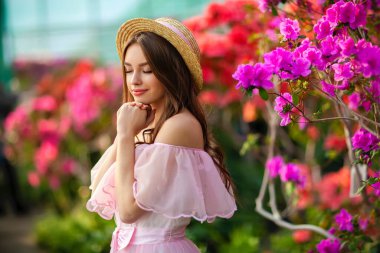 Renkli çiçeklerin yanında duran pembe klasik elbiseli ve hasır şapkalı güzel kız. Romantik bir kadının sanat eseri. Sevimli bir model, poz veriyor ve kameraya bakıyor..