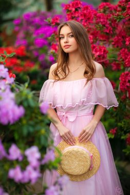 Renkli çiçeklerin yanında duran pembe klasik elbiseli ve hasır şapkalı güzel kız. Romantik bir kadının sanat eseri. Sevimli bir model, poz veriyor ve kameraya bakıyor..