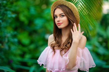 Yeşil arka planda çiçekli pembe bir elbise ve hasır şapkayla duran güzel bir kız. Romantik bir kadının sanat eseri. Sevimli bir model, poz veriyor ve kameraya bakıyor..