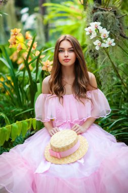 Renkli çiçeklerin yanında duran pembe klasik elbiseli ve hasır şapkalı güzel kız. Romantik bir kadının sanat eseri. Sevimli bir model, poz veriyor ve kameraya bakıyor..