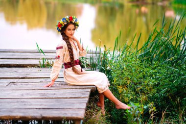 Beautiful long braided hair girl in Ukrainian traditional dress and wreath posing on green nature near river. Portrait of young attractive stylish woman on colorful warm background. clipart