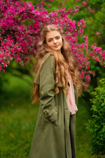 美丽的金发姑娘 卷曲长发 穿着老式花边衬衫和绿色斗篷 站在靠近粉色花朵的春天公园里 温柔浪漫的模特摆出姿势 看着镜头 — 图库照片
