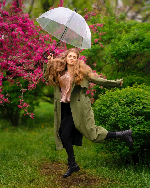 美丽的金发姑娘 卷曲长发 穿着老式花边衬衫和绿色斗篷 站在靠近粉色花朵的春天公园里 轻柔浪漫的造型与透明的伞 — 图库照片