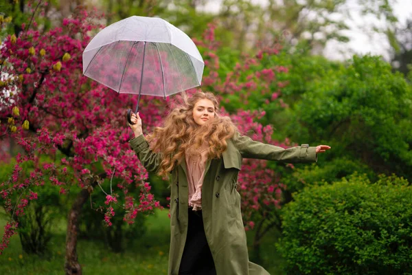 美丽的金发姑娘 卷曲长发 穿着老式花边衬衫和绿色斗篷 站在靠近粉色花朵的春天公园里 轻柔浪漫的造型与透明的伞 — 图库照片